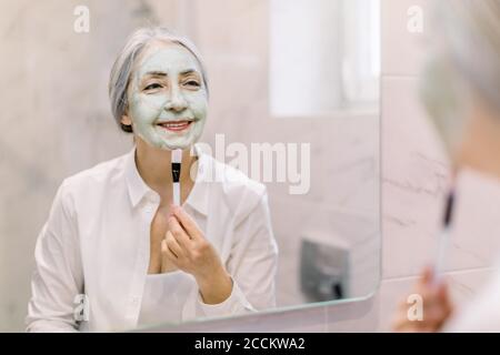 Femme sénior aux cheveux gris, regardant dans le miroir, tenant la brosse cosmétique et appliquant le masque en argile verte pour les soins de la peau du visage dans la salle de bains. Beauté de la peau Banque D'Images