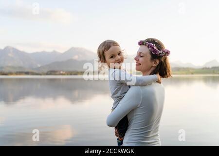 Mère souriante portant une tiara portant sa fille tout en se tenant contre le lac au coucher du soleil Banque D'Images