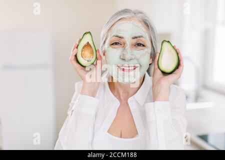 Concept beauté, antirides et soin de la peau. Gros plan de la femme heureuse et belle-look senior à cheveux gris en chemise blanche, avec masque facial en argile à la boue, montrant Banque D'Images