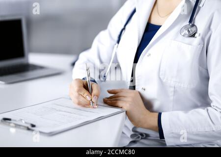 Une femme-médecin inconnue prescrit des médicaments à son patient, à l'aide d'un presse-papiers, tout en étant assise sur le bureau de son cabinet. Femme médecin Banque D'Images