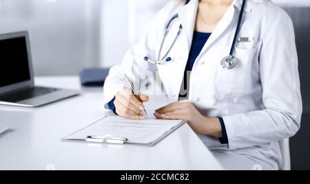 Une femme-médecin inconnue prescrit des médicaments à son patient, à l'aide d'un presse-papiers, tout en étant assise sur le bureau de son cabinet. Femme médecin Banque D'Images