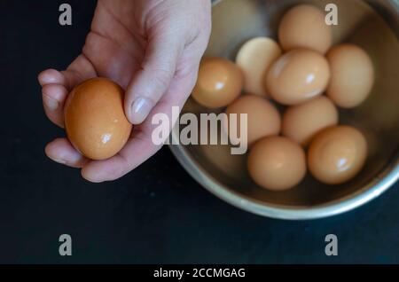 La main mâle contient l'œuf de poulet brun humide. Servir les œufs durs dans un bol en métal avec de l'eau à côté. Processus de préparation du déjeuner. Vue de dessus inclinée. Banque D'Images