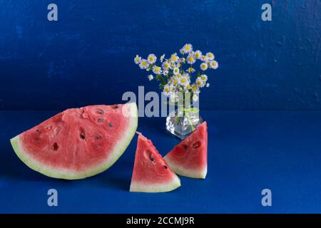 Trois tranches de melon d'eau frais mûr reposent sur un fond bleu foncé. Dans un vase en verre, un bouquet d'Erigeron à petits feuilles. Gros plan Banque D'Images