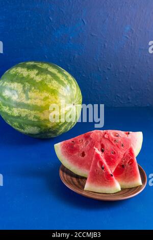 Trois tranches de pastèque fraîche mûre sont dans une assiette en argile sur fond bleu foncé. Theres un melon d'eau entier dans le fond. Verticale Banque D'Images