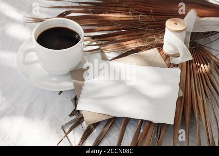 Maquette de papeterie de mariage d'été. Carte de vœux vierge, enveloppes, ruban en soie et feuille de palmier sèche au soleil. Tasse de café sur table blanche Banque D'Images