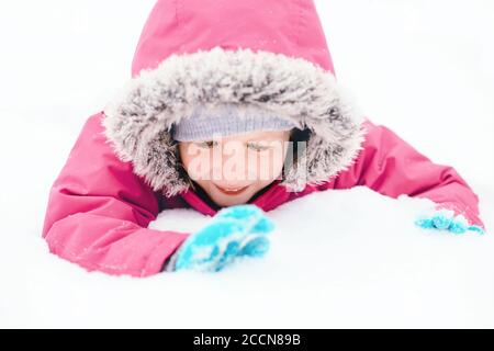 Drôle caucasien souriant fille dans chaud hiver vêtements rose veste jouant avec la neige. Enfant mignon couché sur le sol pendant la journée froide d'hiver enneigée à la chute de neige Banque D'Images