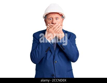 Senior beau gris-cheveux homme portant le costume et l'architecte hardhat choqué couvrant la bouche avec des mains pour erreur. Concept secret. Banque D'Images