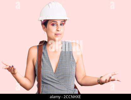 Jeune belle femme portant le casque d'architecte et sac en cuir expression sans indice et confuse avec les bras et les mains soulevées. Doute concept. Banque D'Images