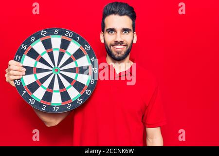 Jeune homme beau avec la barbe tenant le dartboard regardant positif et se tenir debout et sourire avec un sourire confiant montrant les dents Banque D'Images