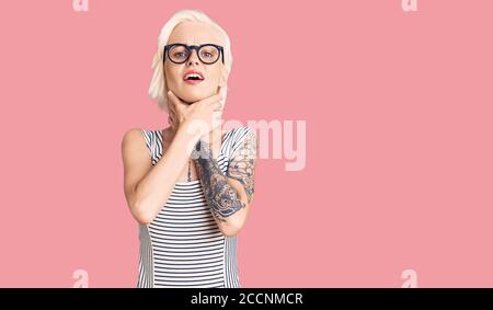 Jeune femme blonde avec tatouage portant des vêtements et des lunettes décontractés criant et suffoquer parce que la cornière douloureuse. Problème de santé. Asphyxiate et suic Banque D'Images