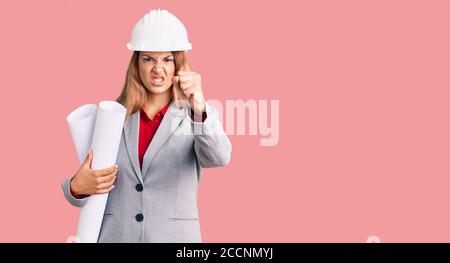 Belle jeune femme portant le casque d'architecte tenant le projet de construction agacé et frustré criant avec la colère, hurlant fou avec la colère et la main ra Banque D'Images