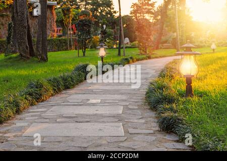 Chemin pavé en pierre dans le parc, au milieu de la pelouse verte et des arbres. Les feux de rue s'allument au coucher du soleil. Batumi, Géorgie. Banque D'Images