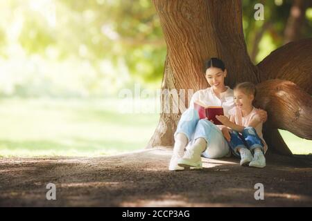 Mère et fille assise sous l'arbre sur la pelouse d'été. Bonne famille jouant à l'extérieur. Jolie jeune maman lisant un livre à son enfant dans le parc Banque D'Images