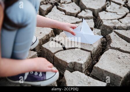 La fille abaisse le bateau en papier sur le sol sec et fissuré. Crise de l'eau et changement climatique concept. Réchauffement de la planète Banque D'Images