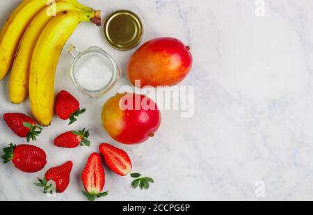 Ingrédients pour préparer des smoothies ou des cocktails : fraise, mangue, banane. Baies, fruits et verre (pot Mason) sur une table en marbre. Concept de santé Banque D'Images