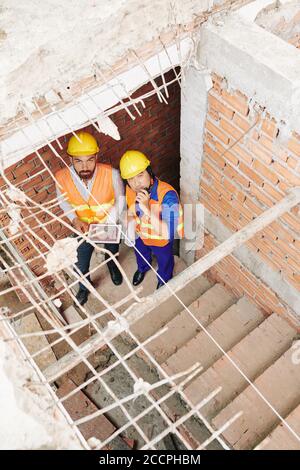 Les constructeurs avec une tablette informatique se baladant dans les escaliers du bâtiment en construction, parlant sur walkie-talkie et regardant vers le haut Banque D'Images