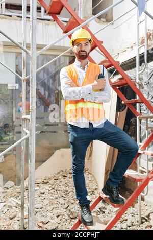 Portrait de l'entrepreneur confiant heureux dans la veste orange vif et hardhat debout sur une échelle dans un bâtiment en construction avec talkie-walkie entre les mains Banque D'Images