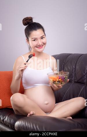 Nutrition enceinte concept sain. Bonne jeune belle femme enceinte asiatique mange de la salade du bol à salade sur le canapé avec le visage smiley. Soins de santé avec RE Banque D'Images