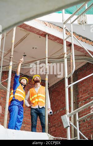 Constructeurs multiethniques en gilets orange debout à l'étage supérieur de construction et discussion sur la rénovation du toit Banque D'Images