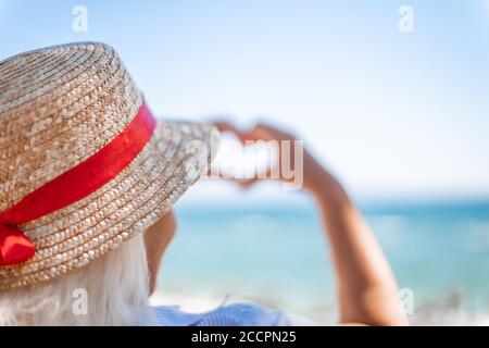 Blonde fille dans un chapeau de paille avec son dos fait signe d'amour avec ses mains sur fond bleu de mer Banque D'Images