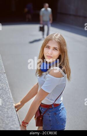 Belle jeune femme caucasienne d'environ 25 ans avec longue les cheveux blonds et les écouteurs sont habillés dans un style décontracté et tendance le tissu marche près de l'uedn Banque D'Images