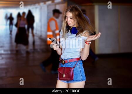 Jeune femme caucasienne attirante d'environ 25 ans avec longue cheveux blonds et écouteurs et habillés en tissu tendance est en passe-bas avec Banque D'Images