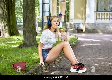 Belle jeune fille caucasienne environ 25 ans avec des cheveux blonds mange de la crème glacée dans le parc public. Femme blanche en tissu tendance avec plate-forme Banque D'Images