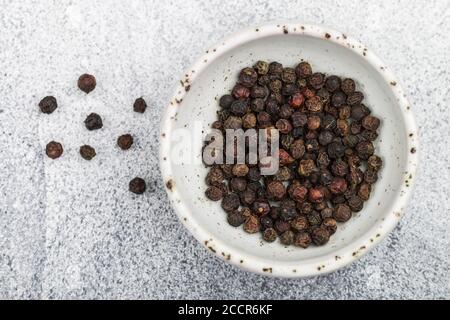 Grains de poivre noir biologique séchés dans un bol blanc sur fond de béton léger ou de pierre. Épices et assaisonnements. Mise au point sélective, vue de dessus et Banque D'Images