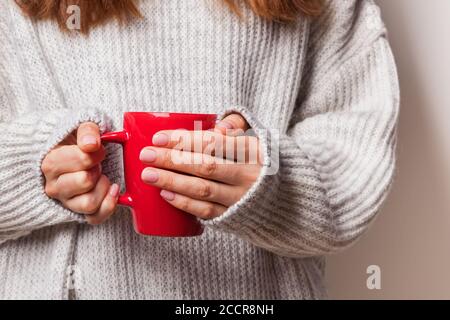 Gros plan des mains des femmes dans un chandail tricoté tenant une tasse rouge de boisson chaude. Banque D'Images