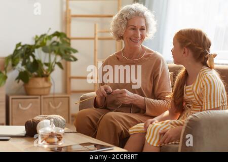 Portrait aux tons chauds d'une femme âgée souriante tricotage avec une jolie fille la regardant dans une maison confortable éclairée par la lumière du soleil, espace de copie Banque D'Images