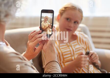 Portrait aux tons chauds d'une femme âgée méconnue qui prend une photo avec un smartphone de mignonne fille aux cheveux rouges dans un intérieur confortable Banque D'Images