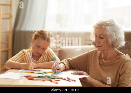 Portrait ton chaud d'une femme âgée souriante gardiennant mignon rouge une fille aux cheveux et un dessin ensemble tout en étant assis près d'une table basse dans le salon confortable Banque D'Images