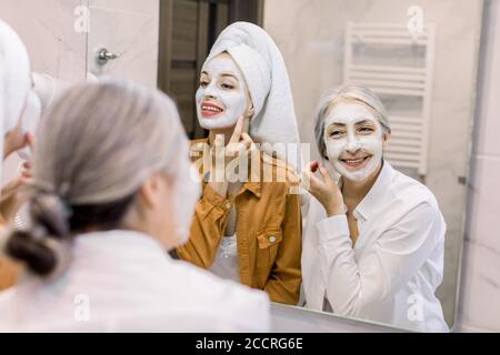 Spa familial heureux. Réflexion dans le miroir de deux femmes charmantes, jeune fille avec une serviette sur la tête et sa grand-mère senior, appliquant un masque facial de boue Banque D'Images