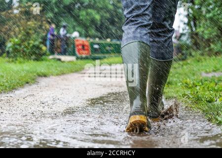 Vue rapprochée de l'arrière des bottes en caoutchouc vertes portées par une femme qui éclate dans des flaques sous une forte pluie d'été britannique, en marchant le long du chemin de halage du canal britannique. Banque D'Images