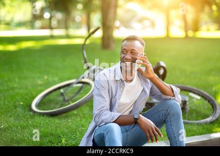 Beau noir homme assis sur le sol près de son vélo et parler au téléphone portable au parc Banque D'Images