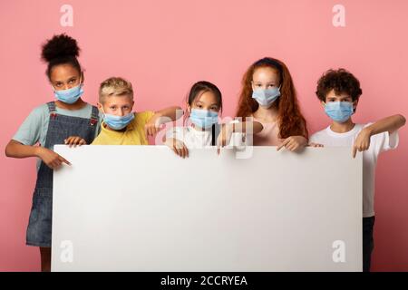 Groupe multiethnique d'enfants dans des masques médicaux pointant sur une bannière vierge sur fond rose, maquette pour le design Banque D'Images