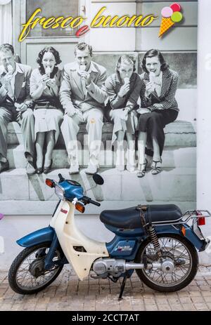 un mobylette ou une moto sur une rue grecque garée au bord de la route devant une affiche représentant les jeunes assis. Banque D'Images