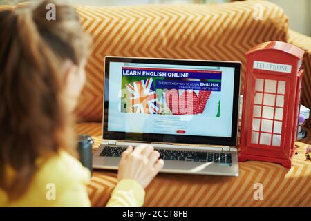 Vue de derrière une femme dans la maison en journée ensoleillée apprenant la langue anglaise en ligne sur un ordinateur portable. Banque D'Images