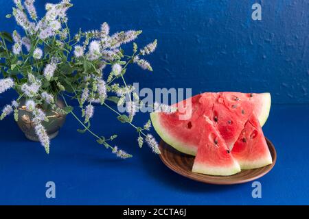 trois tranches de pastèque fraîche mûre sont dans une assiette en argile sur fond bleu foncé. Dans un vase en verre, un bouquet d'origan. Gros plan Banque D'Images