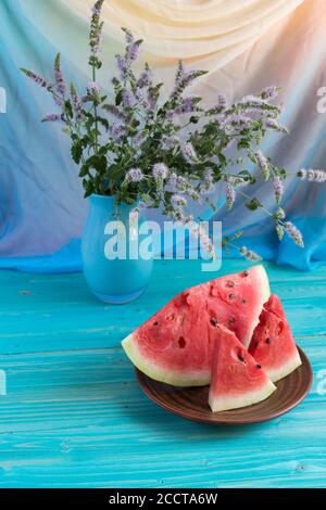 Trois tranches de pastèque fraîche mûre sont dans une assiette en argile sur la table bleue près de la fenêtre. Dans un vase, un bouquet d'origan. Verticale Banque D'Images