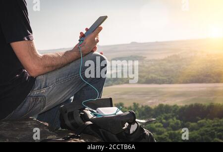 Man On a rando utilise un smartphone tout en se chargeant depuis la banque d'énergie sur le rocher à l'aube. Mode de vie sain et communication Banque D'Images