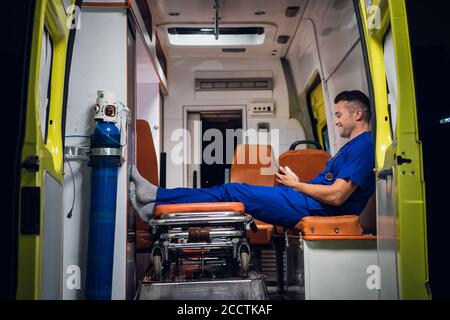 Jeune stagiaire dans un uniforme bleu se reposant et parcourant son téléphone dans la voiture d'ambulance. Banque D'Images
