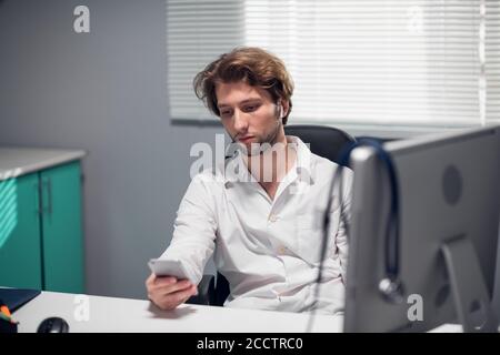 Un médecin fatigué avec le casque sans fil pour vérifier ses messages vocaux sur le téléphone. Banque D'Images