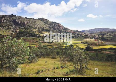 Paysage typique de Madagascar dans la région près de Tsiafahy, de petites collines couvertes d'herbe verte et de buissons, des maisons en argile rouge et des champs de riz près. Banque D'Images