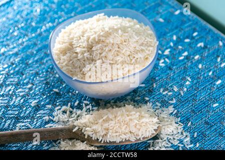 Riz pelé et transformé dans un petit bol. Pelle en bois avec riz sur fond bleu. Nourriture humaine. Céréales prêtes à consommer. Banque D'Images
