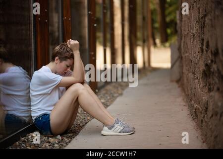 Une fille bien réfléchie et triste. Portrait d'une femme avec des cheveux courts regardant vers le bas tristement pensant assis sur le sol à l'extérieur à côté d'un mur de verre backgrou Banque D'Images