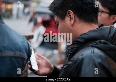 Gros plan jeune homme asiatique porte une capuche noire et un diamant ecouteurs lorsque vous utilisez un smartphone et que vous voyagez en ville avec des amis Banque D'Images