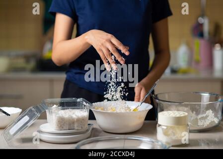 Femme ajoutant de la noix de coco râpée dans la pâte à biscuits tout en faisant maison petits gâteaux dans la cuisine à la maison Banque D'Images