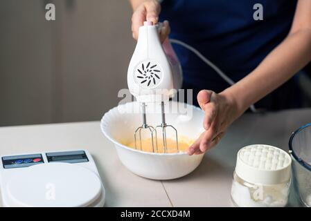 Femme utilisant un batteur électrique pour mélanger les ingrédients pour la pâte de sucre, les oeufs battus, le beurre et la farine tout en faisant des biscuits dans la cuisine à la maison de gros plan Banque D'Images