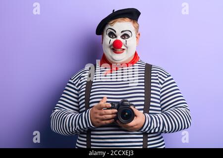 positif impressionnant plump mime avec visage blanc peint et nez clown portant des bretelles et le mouchoir rouge va prendre une photo, isolé arrière-plan bleu Banque D'Images